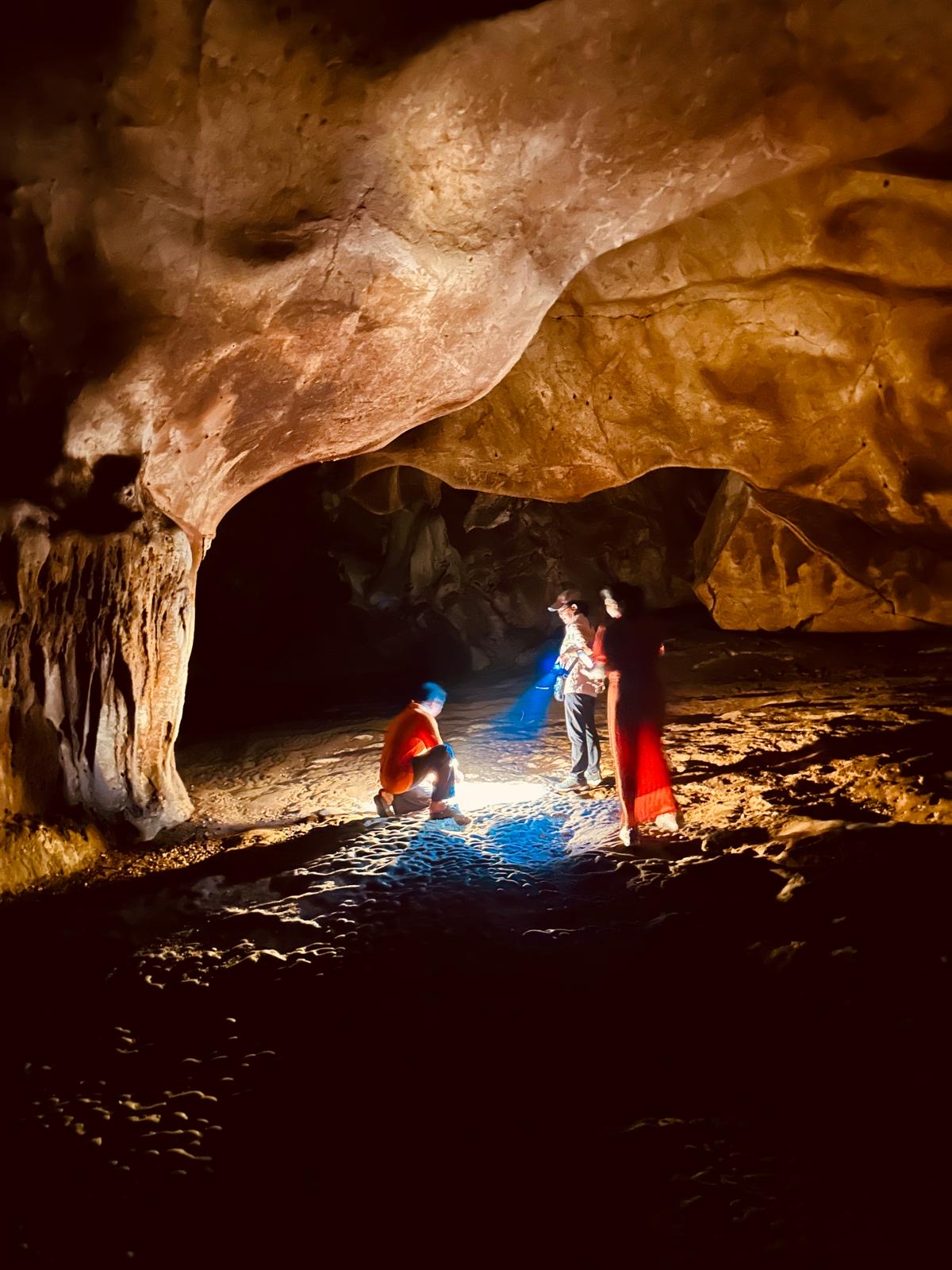 Drei Menschen erkunden mit Lampen eine Höhle in Kambodscha unter einer weit geschwungenen Felsdecke.