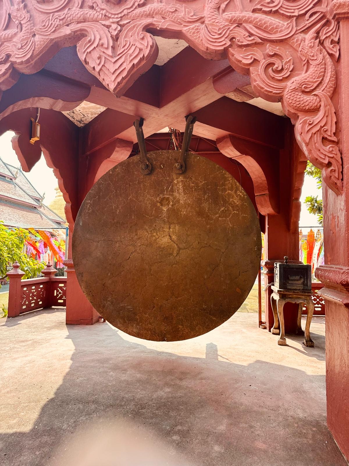 Ein grosser Gong hängt in einem rot geschnitzten Pavillon im Sonnenlicht.