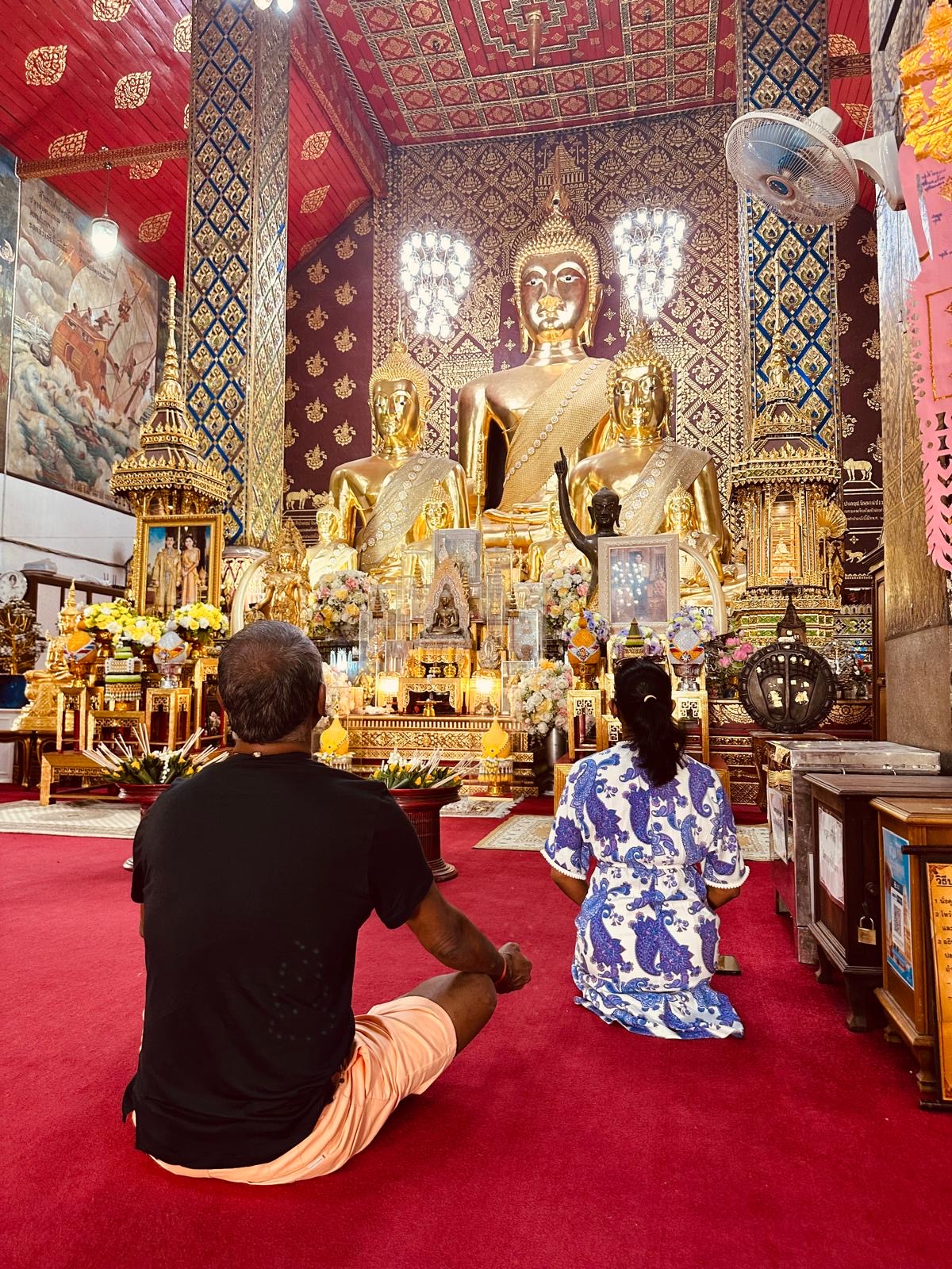 Zwei Menschen sitzen vor goldenen Buddhafiguren im reich geschmückten Tempelraum.